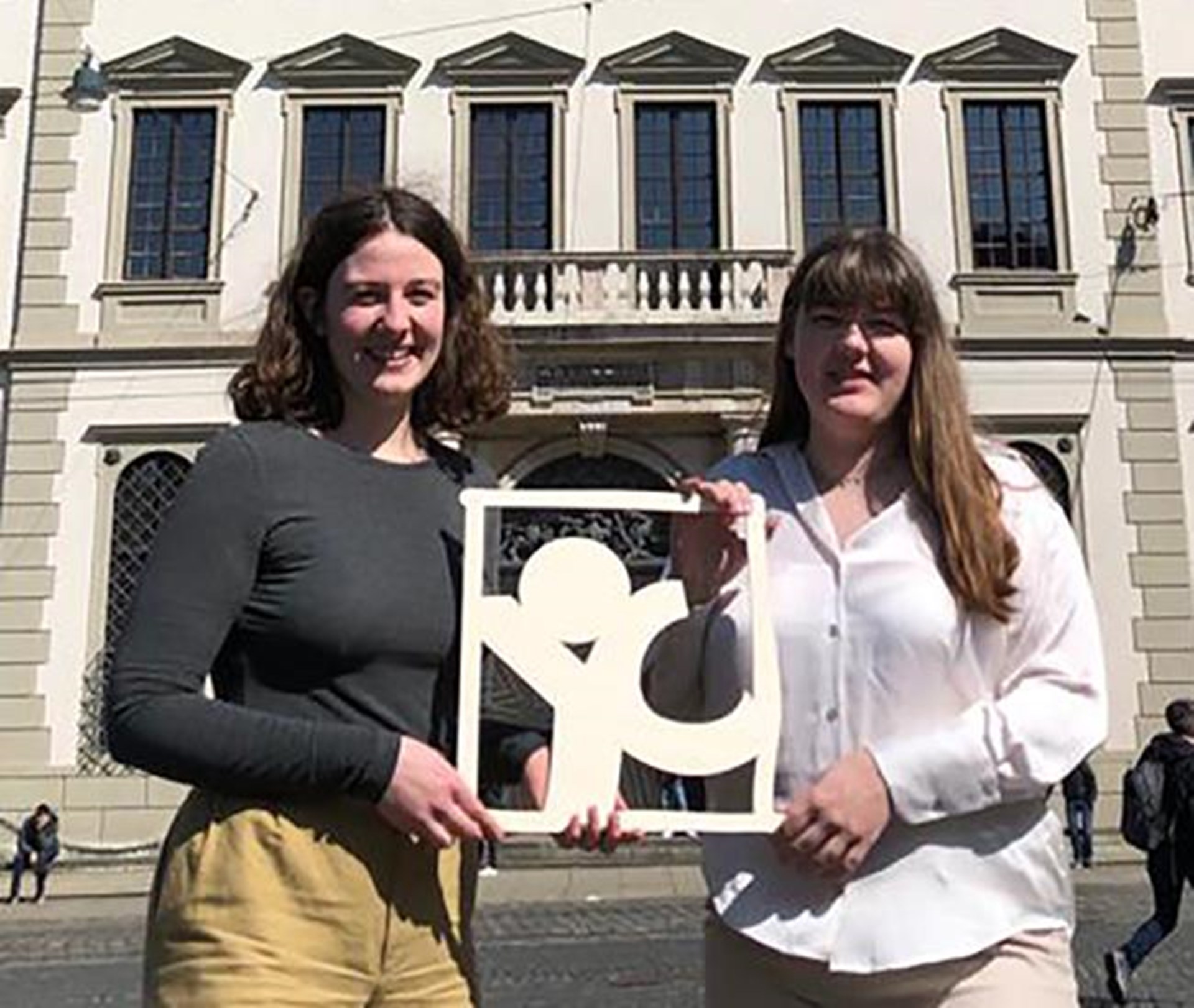 Zwei junge Frauen, die Studentinnen Hannah Schmid und Julia Claesson, stehen vor dem Augsburger Rathaus und werben für young caritas. 